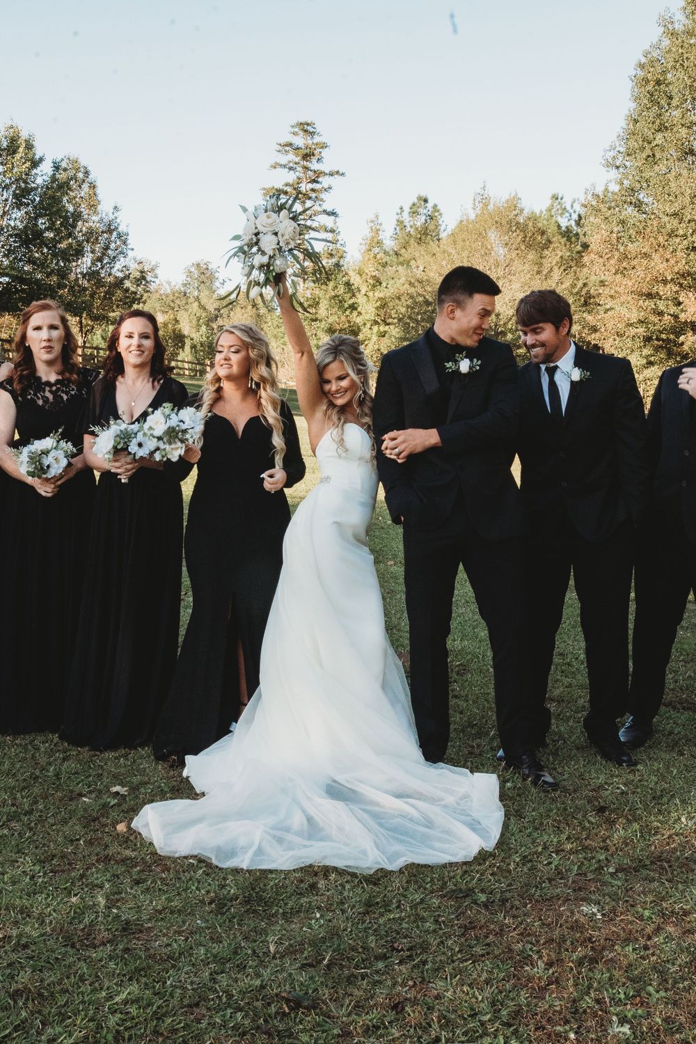 Bride raises bouquet triumphantly with wedding party in black attire on green lawn outdoors.