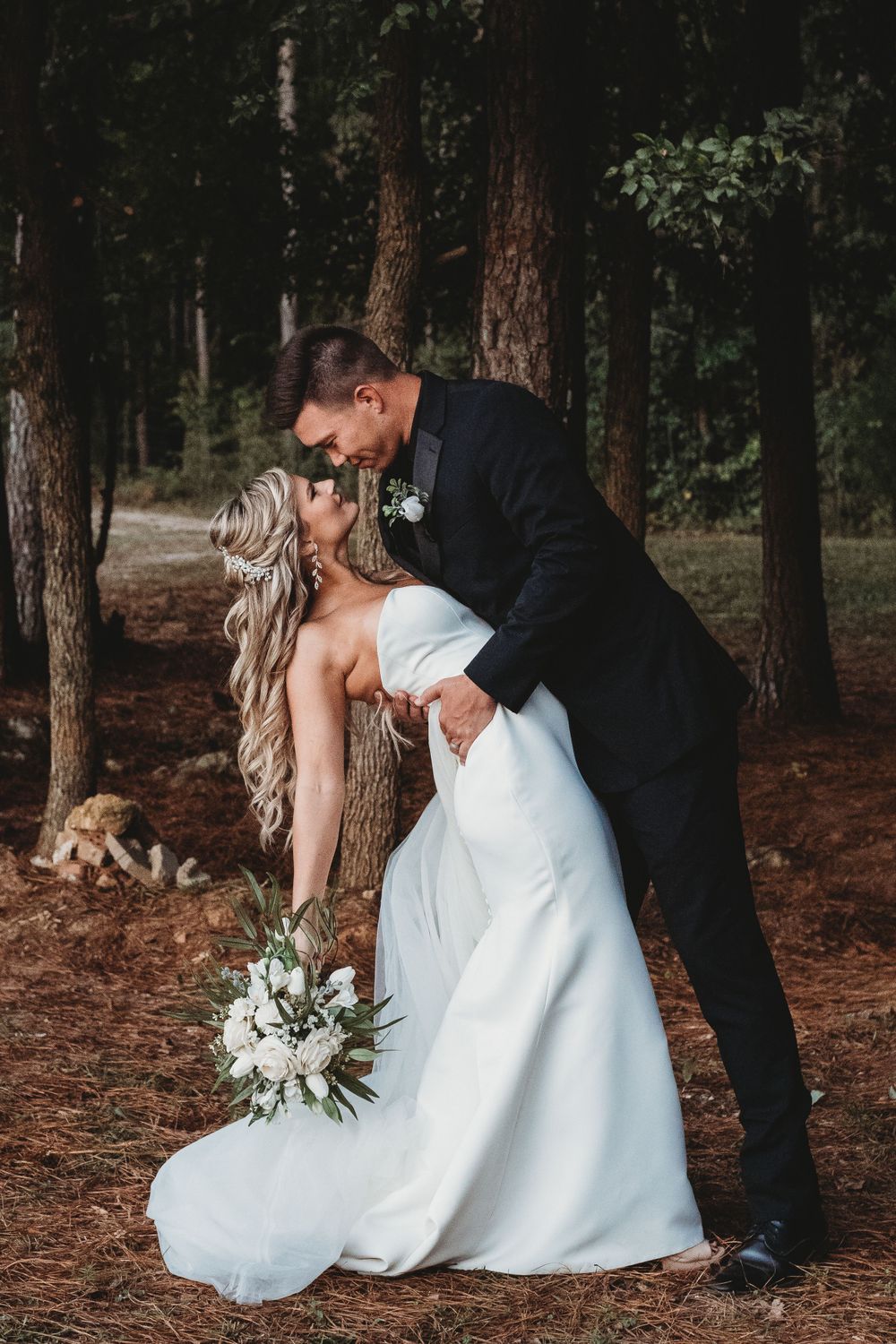 Groom in black suit dipping bride in white gown for a romantic kiss in a pine forest.