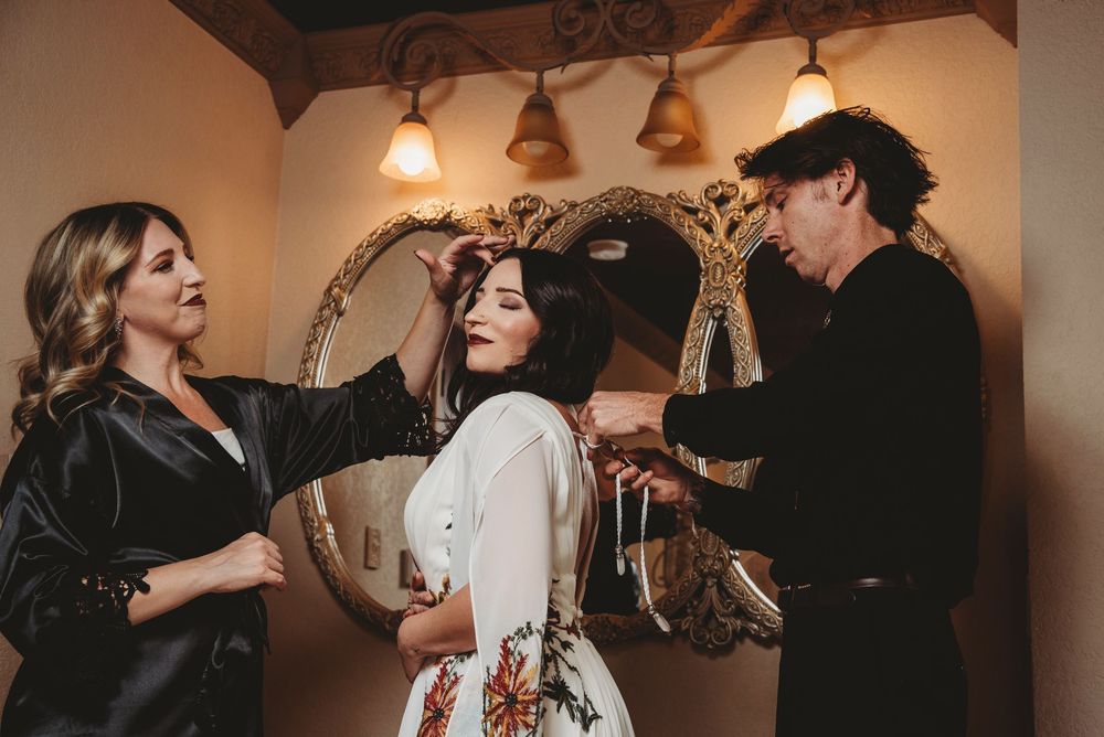 Bride gets hair styled by two attendants in front of ornate gold mirrors before wedding.
