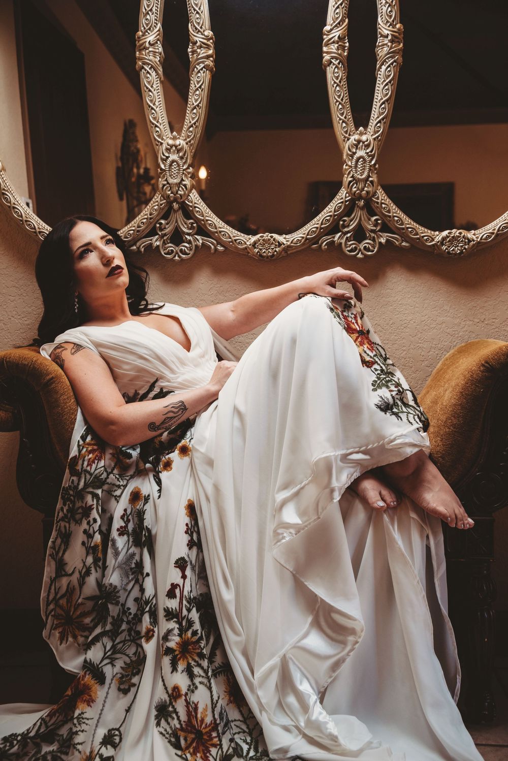 Bride in floral embroidered white wedding dress reclines elegantly on ornate vintage chair indoors.