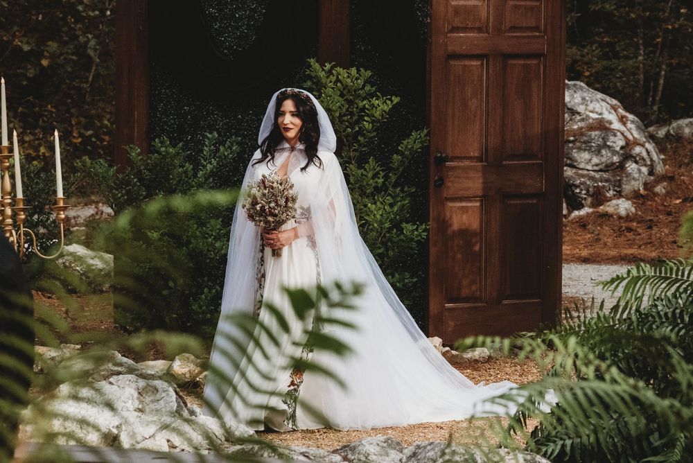 Bride in white gown and veil holds bouquet before rustic wooden doors surrounded by ferns.
