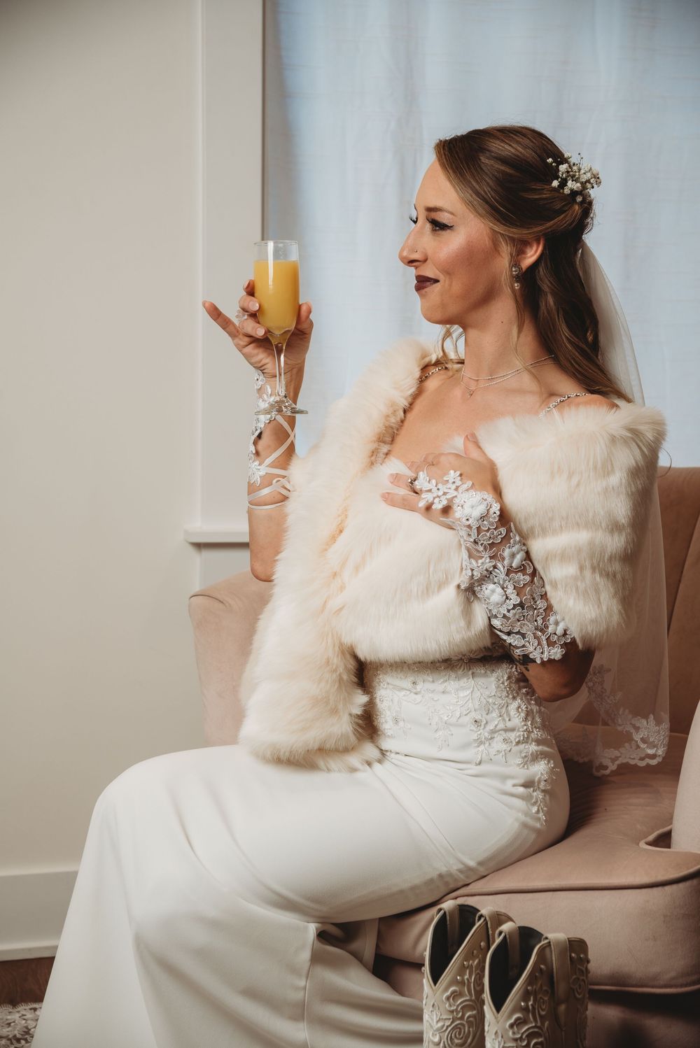 Bride in white lace gown and fur stole sips mimosa while seated, wearing veil and floral hair accessories.