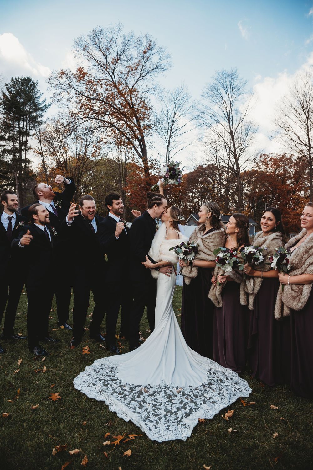Wedding party in burgundy and black attire cheers as bride and groom kiss on autumn lawn.