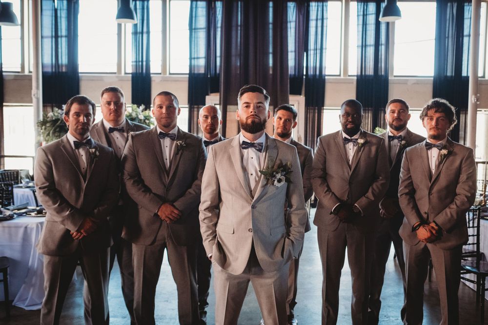 Groom stands with groomsmen all wearing matching grey suits and bow ties indoors.