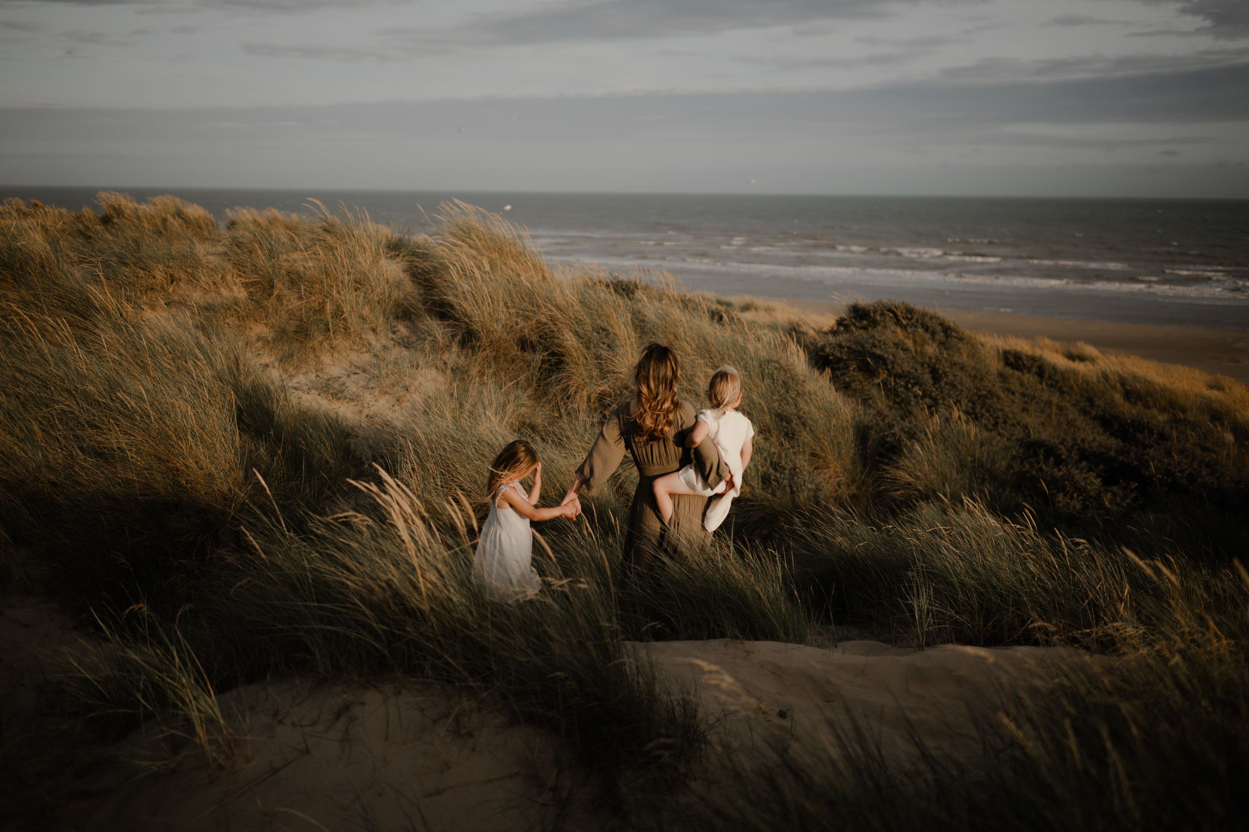 a mother holding hands with her daughter and holding another in her arms is walking down a sand dune at Camber Sands during a photo session