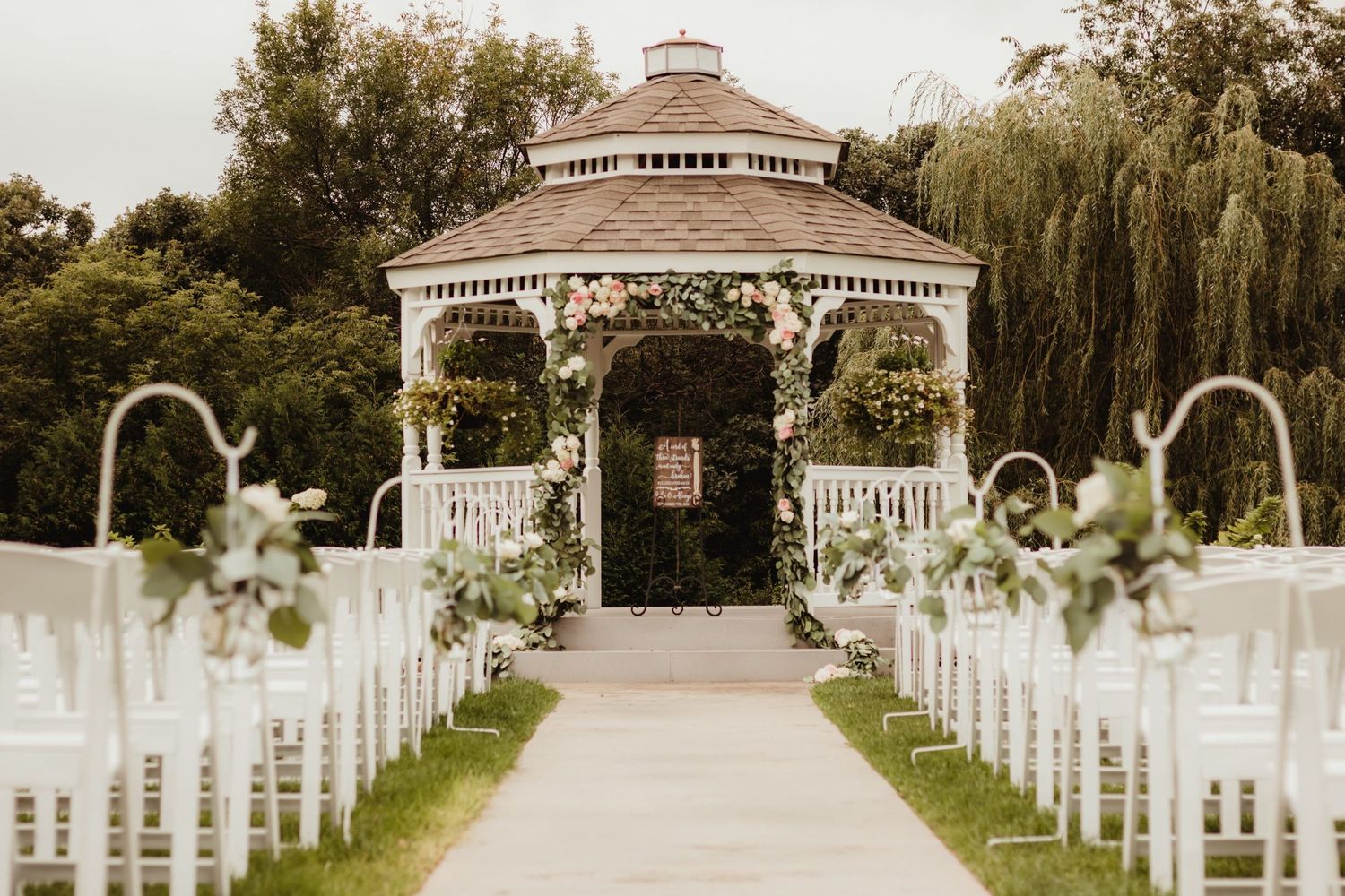 White wedding gazebo with floral arrangements and rows of chairs lining a walkway for an outdoor ceremony.