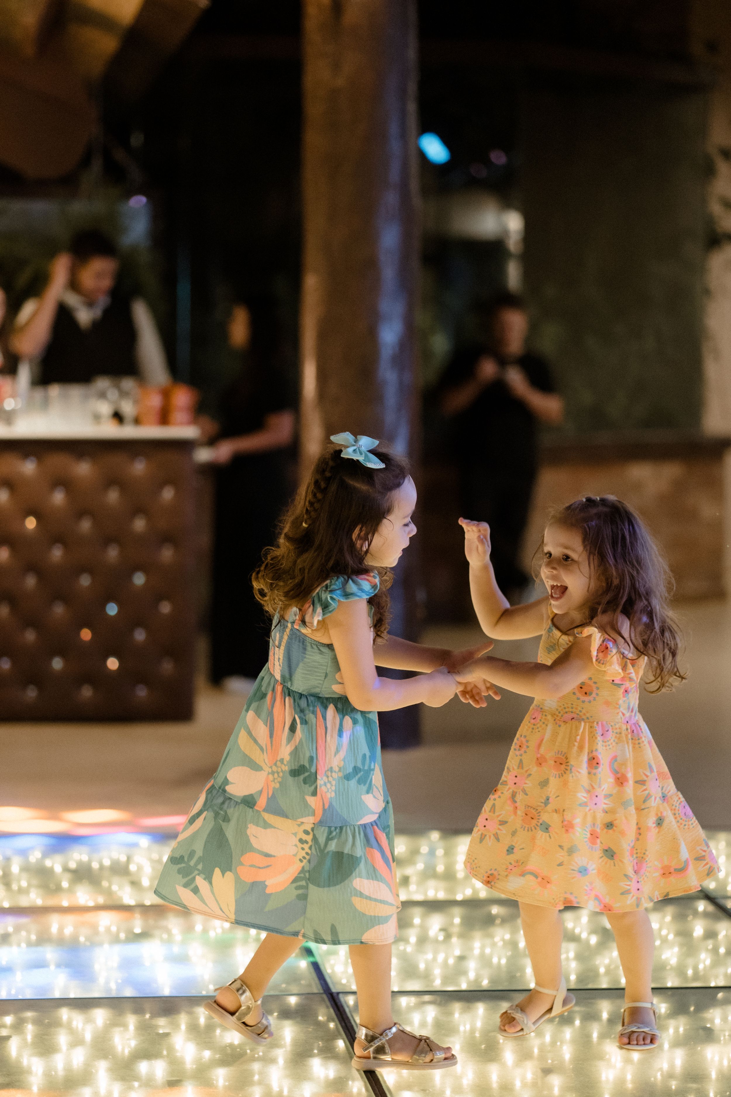 Two young girls in floral dresses dance joyfully on an illuminated dance floor.