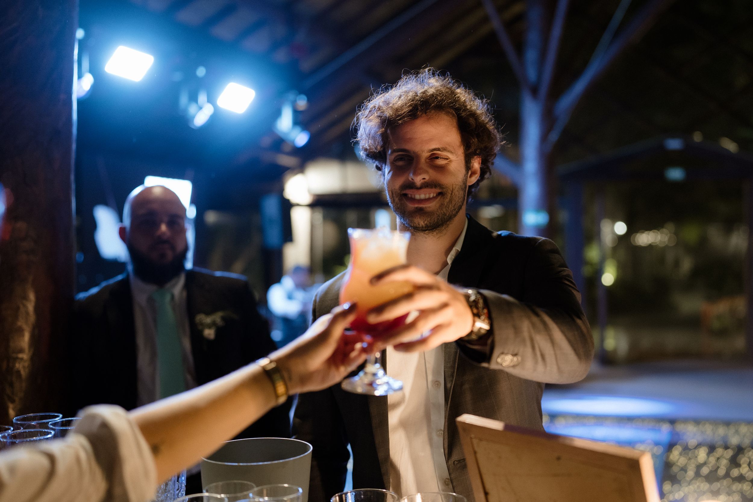 Someone hands a craft cocktail to a guest at an outdoor evening event.