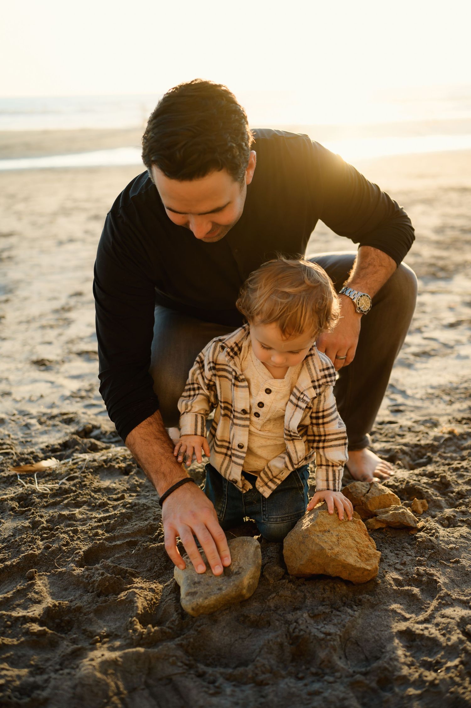 Man and a small child examining a rock on the beach during sunset.
