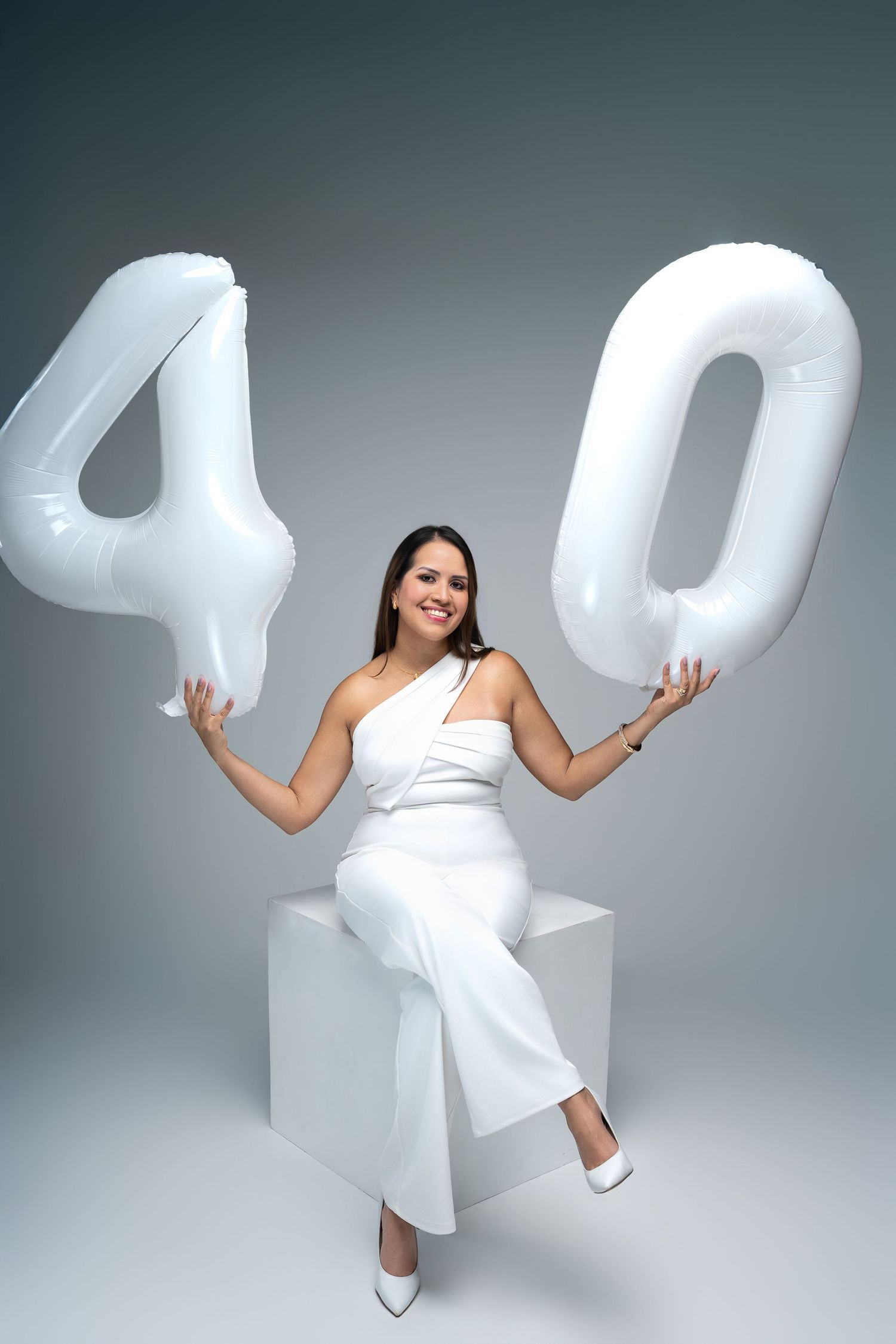 Woman in white jumpsuit sitting on cube, holding large white number 40 balloons, celebrating 40th birthday.