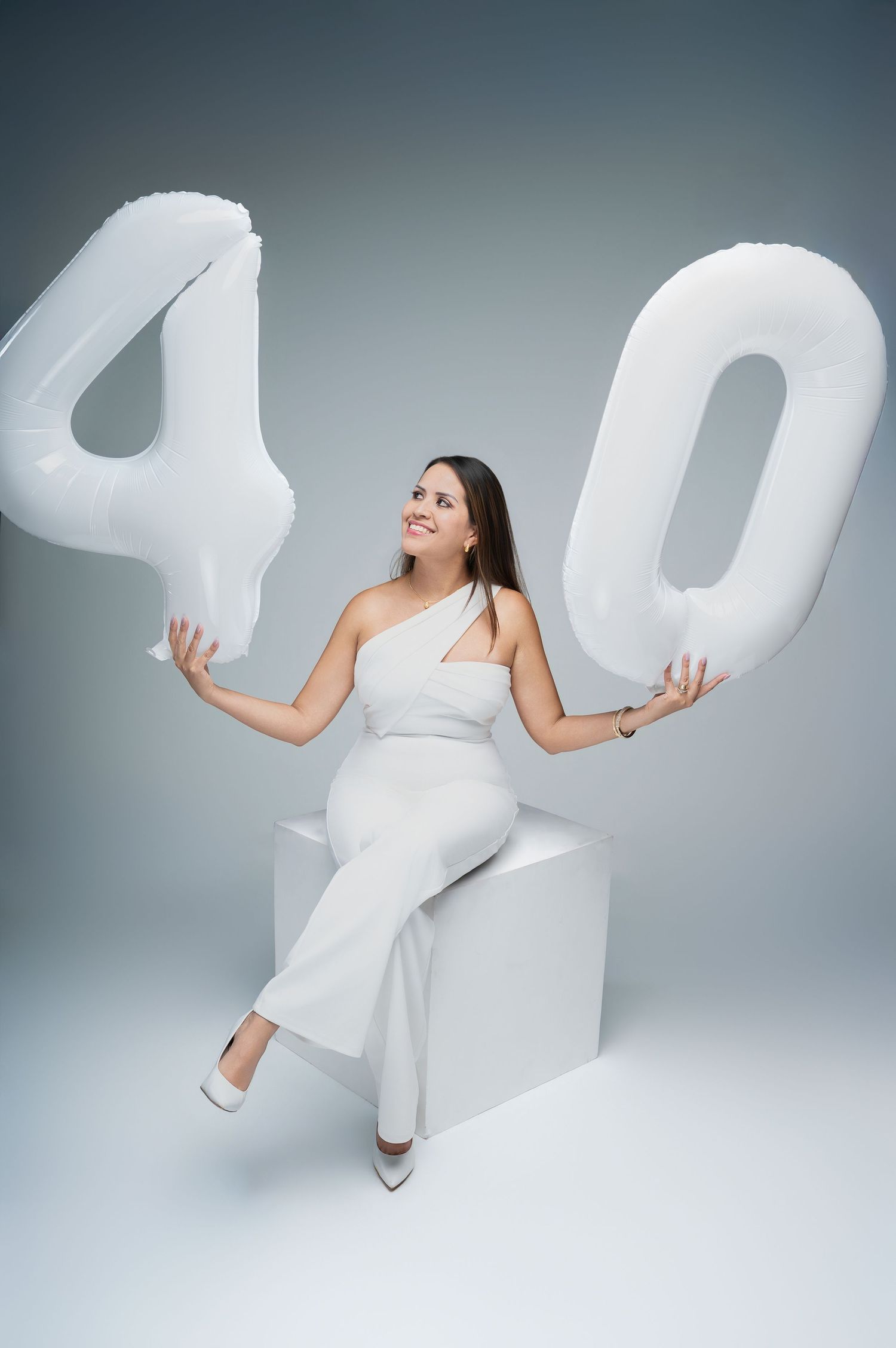Woman in white outfit sitting on cube holding large white balloon number 40 for birthday celebration on gray background.