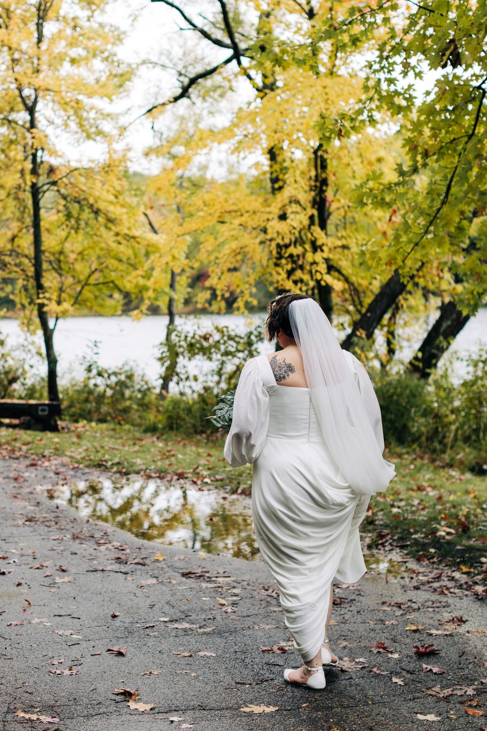 A bride in a white dress walks along a path surrounded by golden fall trees near a lake.