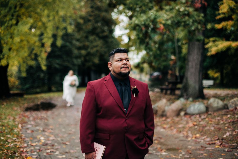 A person in a burgundy suit stands on a leaf-covered path with autumn trees in the background.