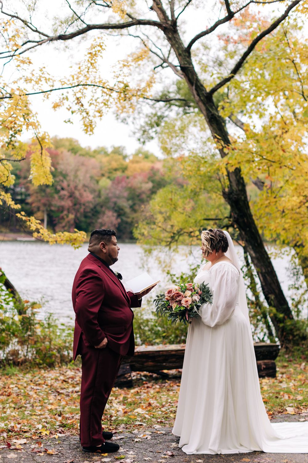 A wedding ceremony taking place beside a lake with vibrant fall foliage in the background.