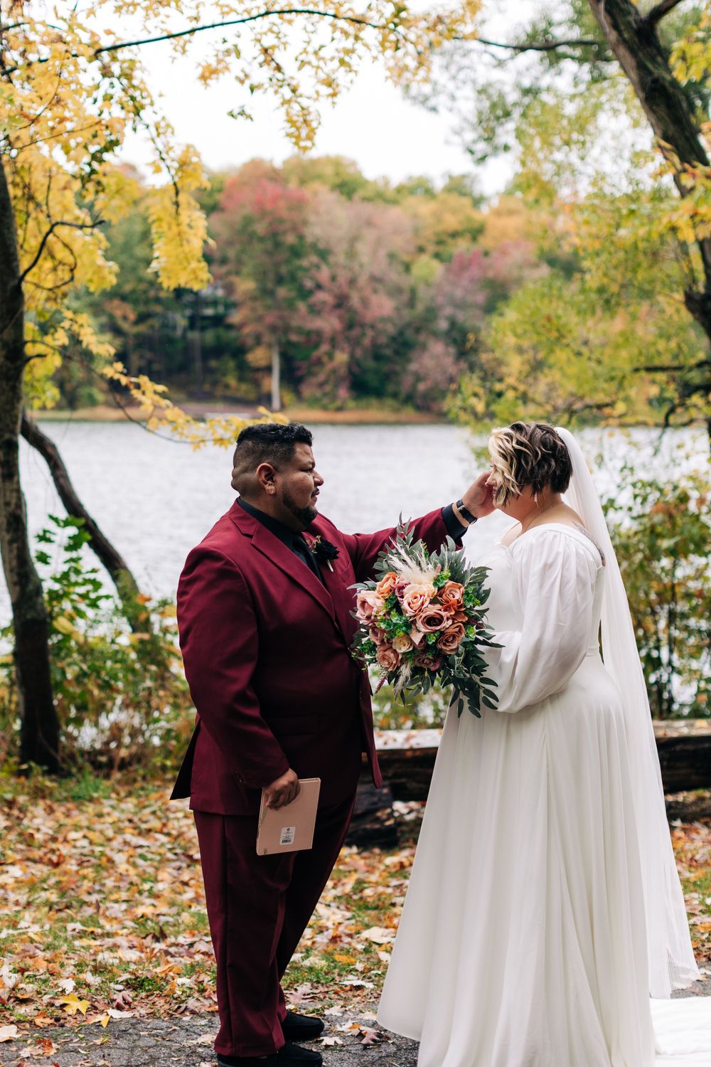 A wedding ceremony takes place alongside a serene lake surrounded by vibrant fall foliage and yellow leaves.