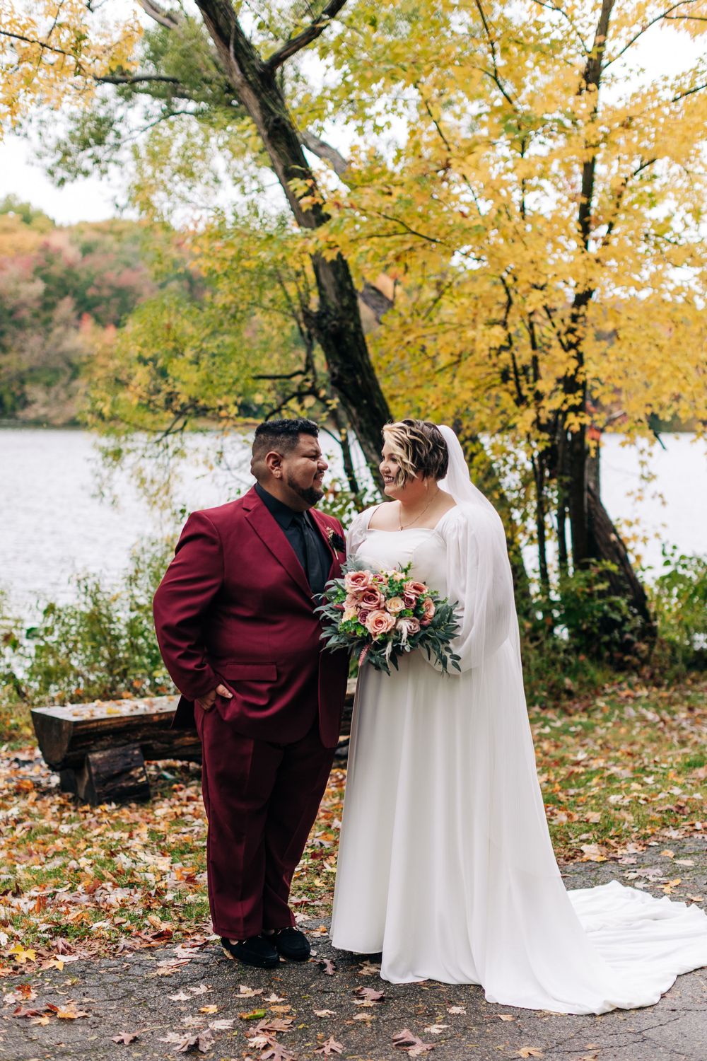 An autumn wedding scene with burgundy and white attire against golden foliage by a lake.