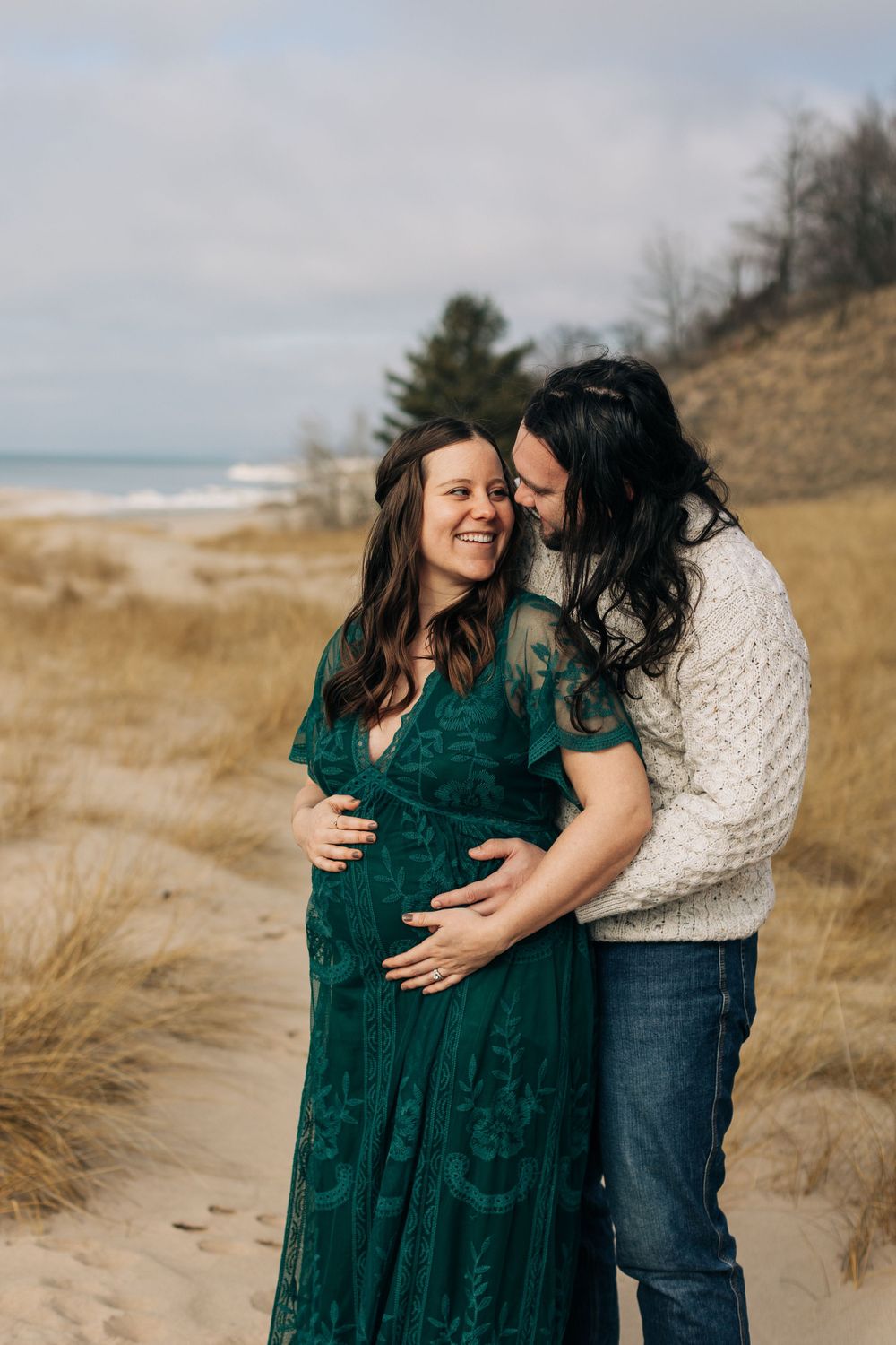 Expectant couple sharing tender moment on beach with sand dunes backdrop.