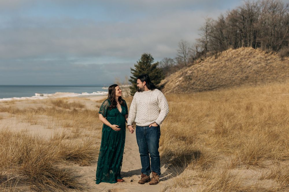 A couple stands together on a sandy beach dune with water and cloudy skies in the background.