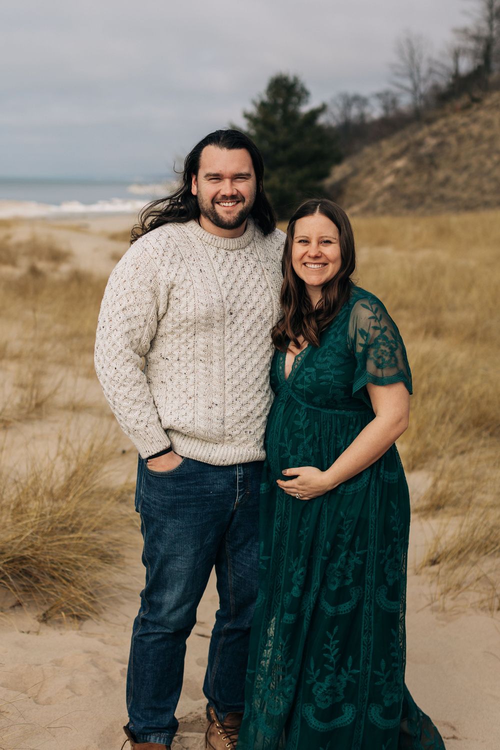Couple in winter attire pose on beach with sand dunes in background.