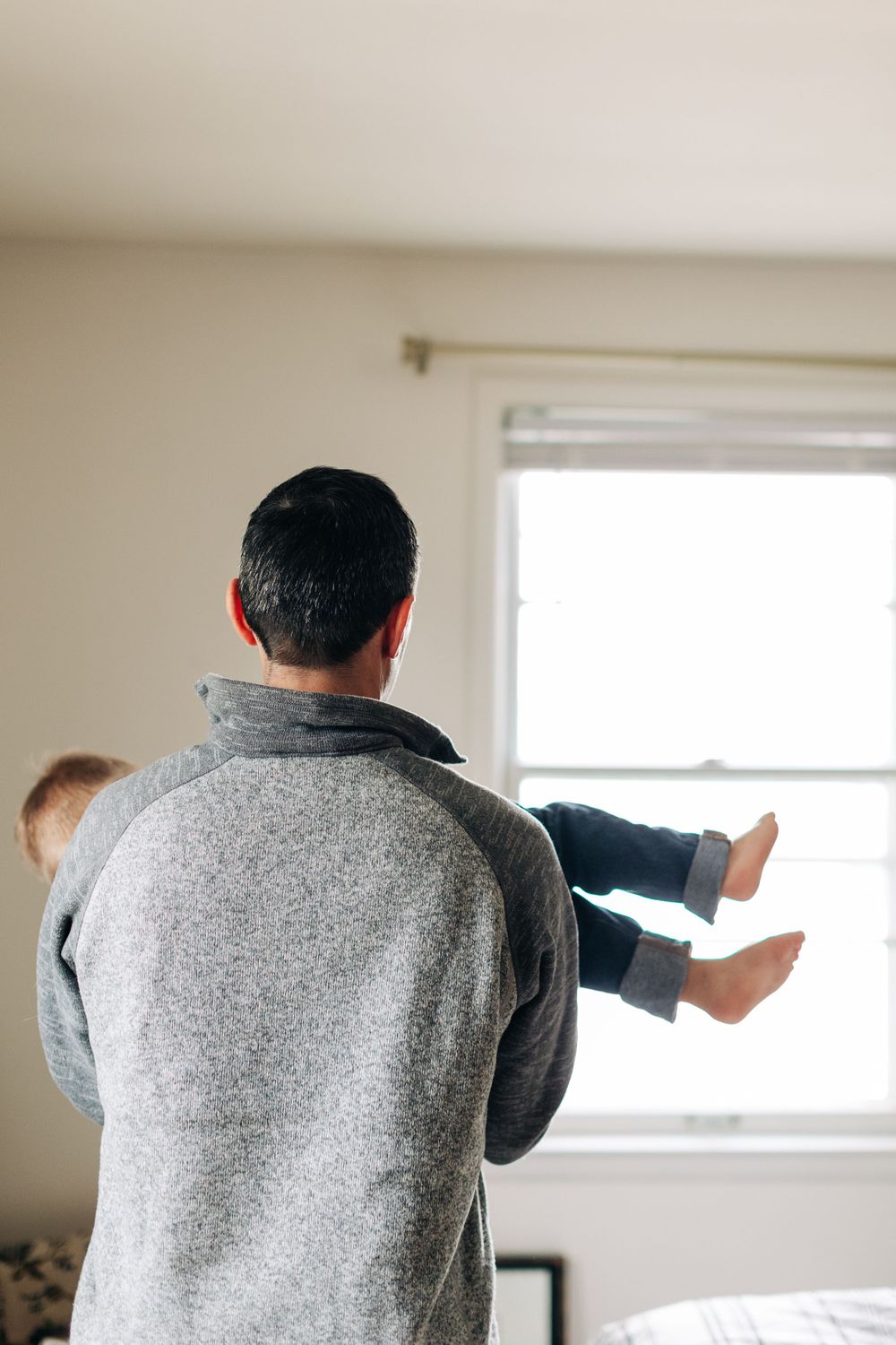 A person wearing a gray sweater holds a child up near a window in a bright room.