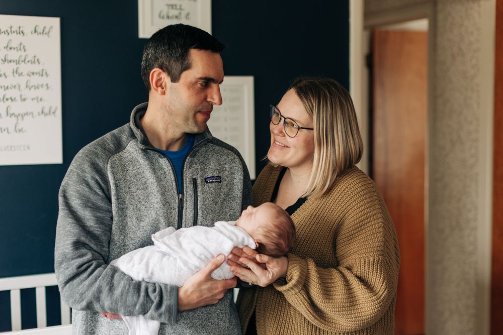 New parents in casual attire holding their newborn in a nursery with dark walls.