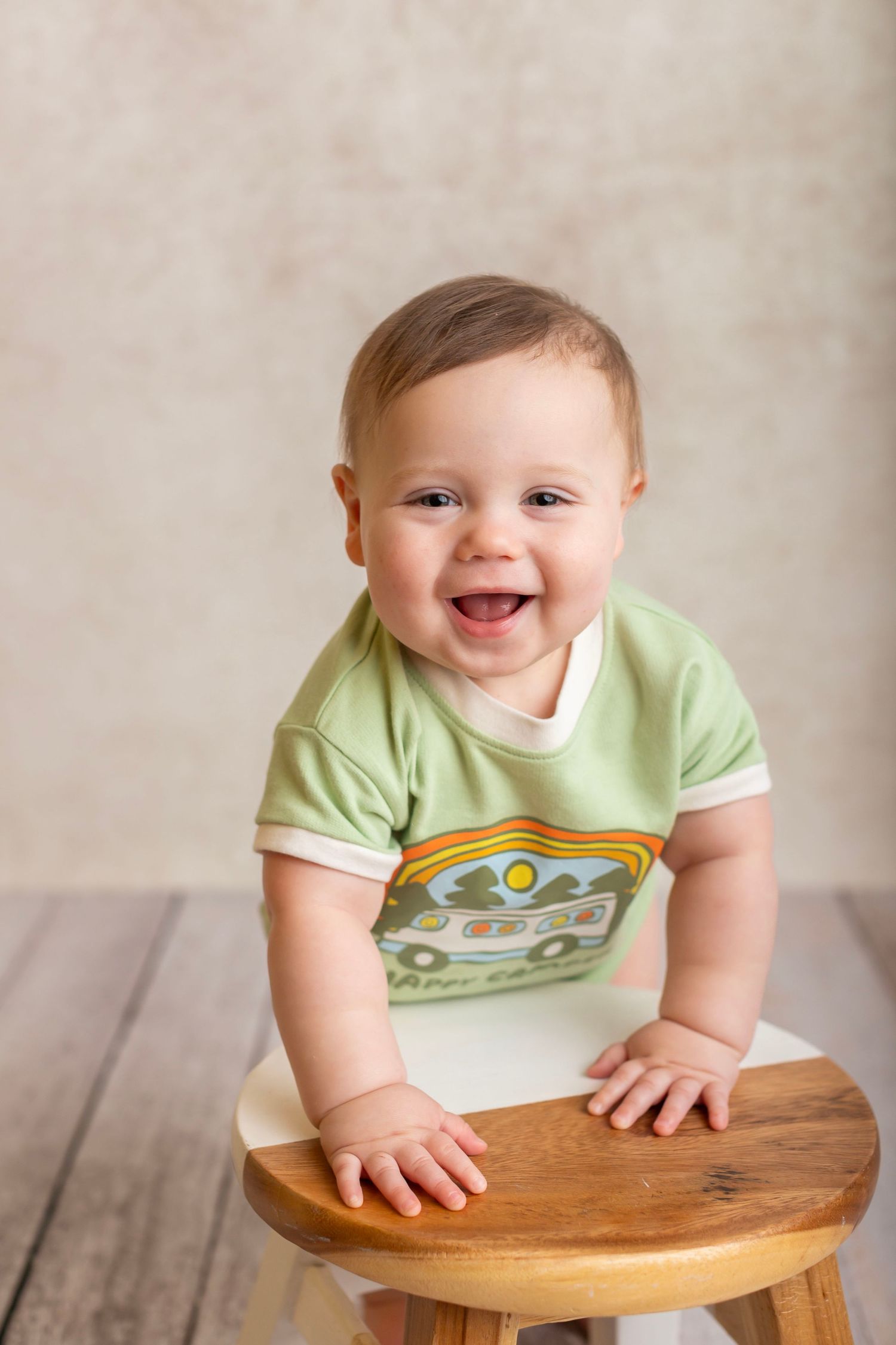 Cheerful baby in light green t-shirt sitting on wooden stool against neutral background.