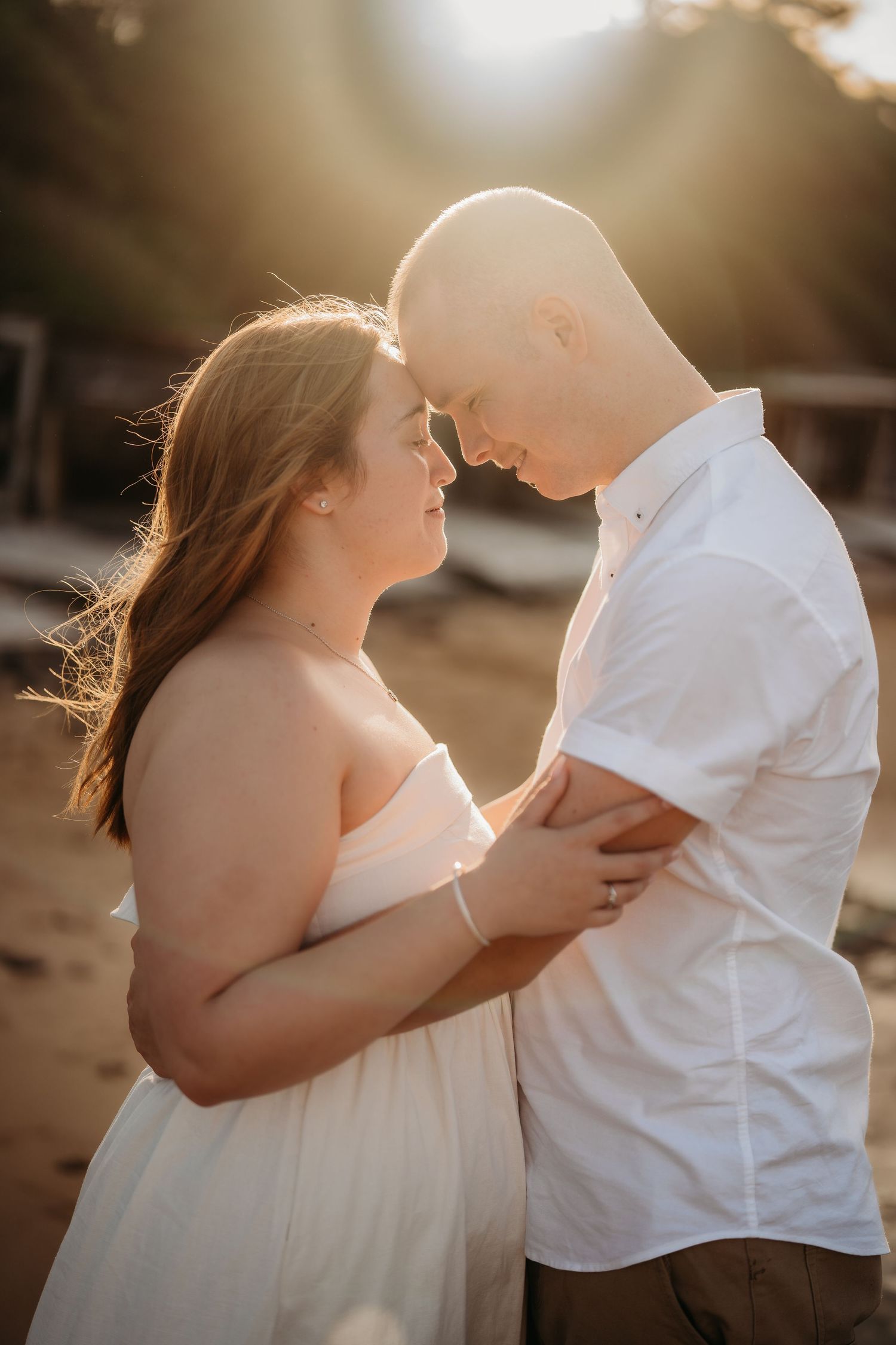 Pregnant couple touching foreheads tenderly in golden backlit beach setting at sunset.