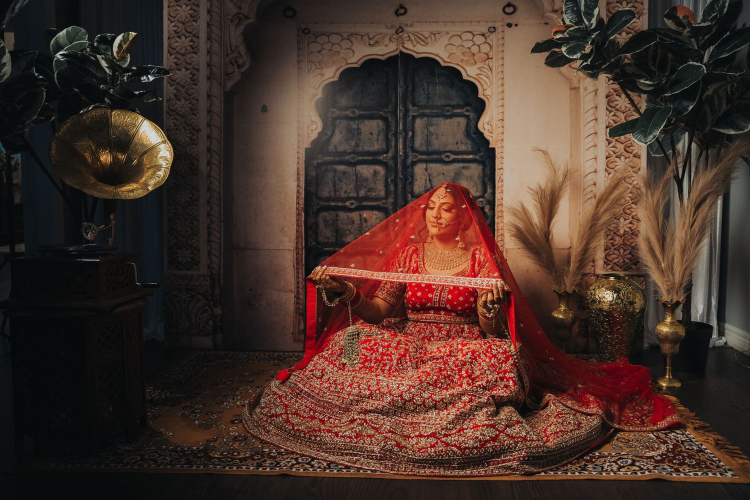 A traditional Indian bridal portrait in a dimly lit room with ornate decor and red lehenga.