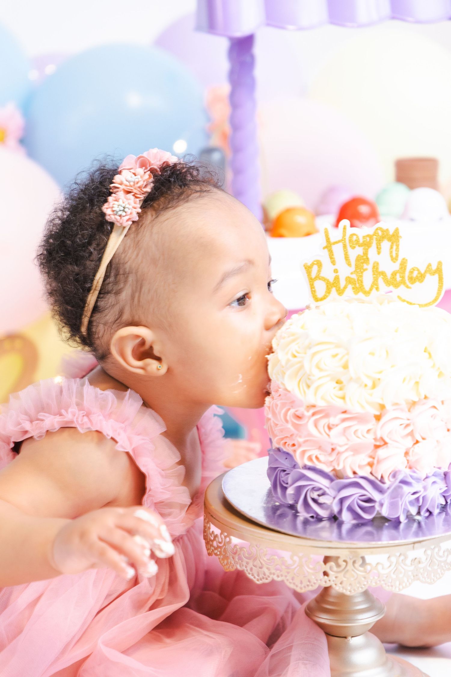 Baby in pink dress celebrates first birthday with elegant purple and pink frosted cake.