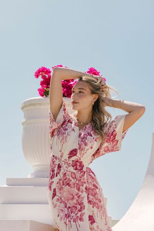 A person in a pink floral dress poses outdoors against a blue sky with white architectural elements and flowers.