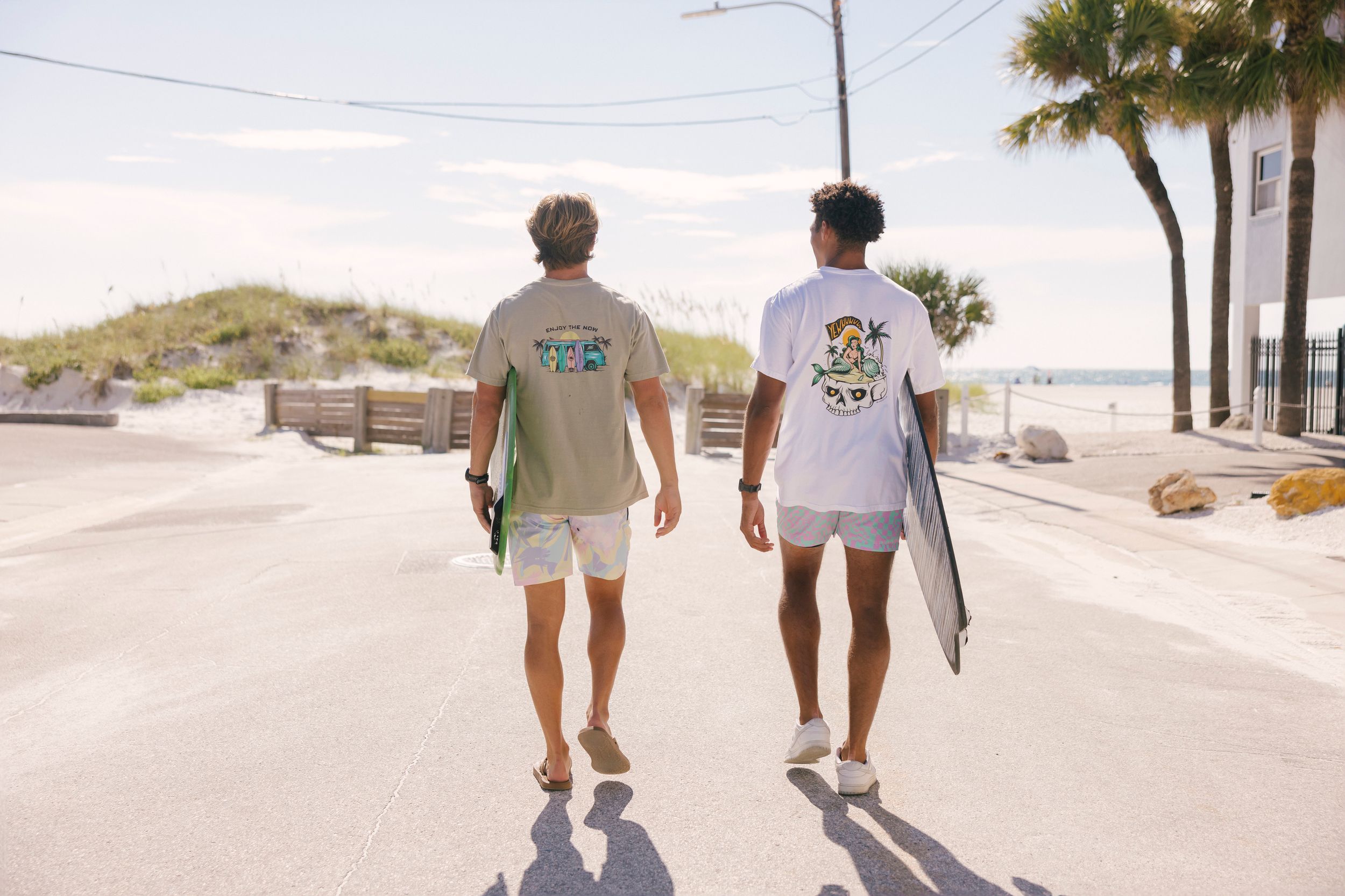 Two surfers walking down a sunny coastal road carrying surfboards on a summer day.