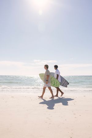 Two surfers carrying their surfboards walk along a sunny white sand beach with ocean waves in the background.