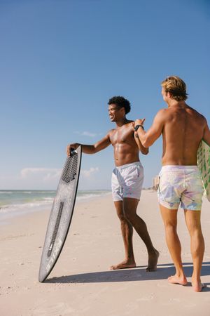 Two surfers in white shorts stand on a sunny beach holding surfboards against a bright blue sky and ocean backdrop.