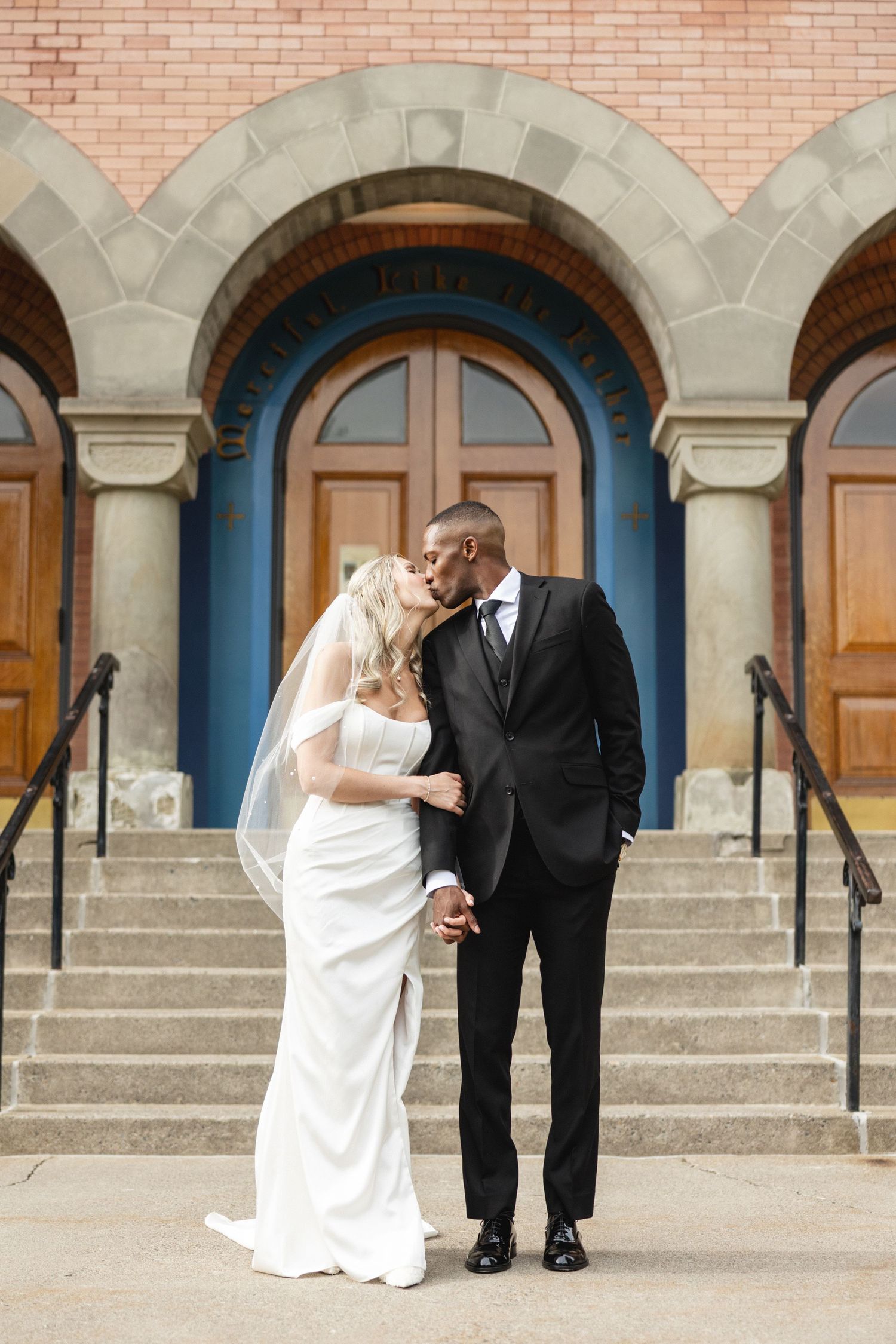 Bride and groom share a kiss on church steps, wearing white gown with veil and black tuxedo.