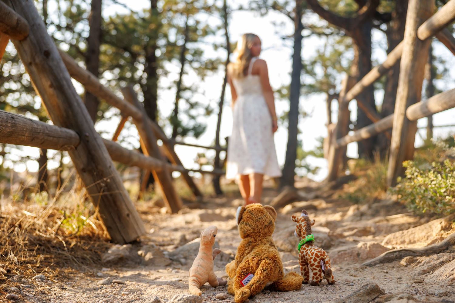 Girl in white dress walks away from stuffed animal toys on a sandy forest path surrounded by trees.