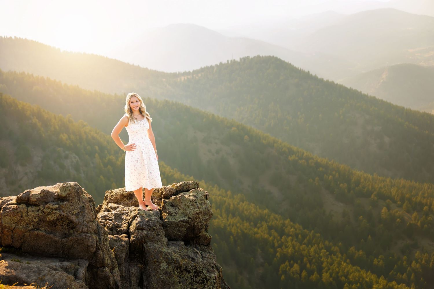 Woman in white dress standing on rocky mountain peak with scenic forested valley and golden sunset behind her.