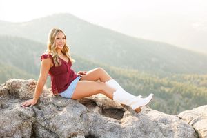 Blonde woman in maroon top and denim shorts sitting on rocky mountain overlook wearing white cowboy boots.