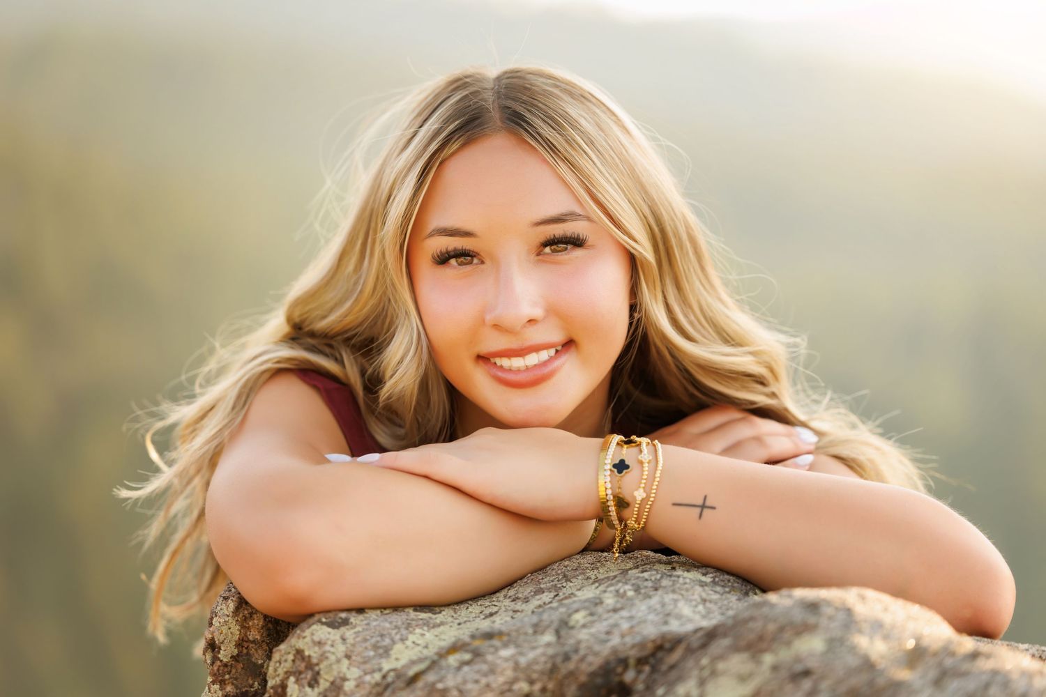 Smiling blonde teenage girl resting on a rock outdoors, wearing bracelets, with a soft bokeh mountain background.