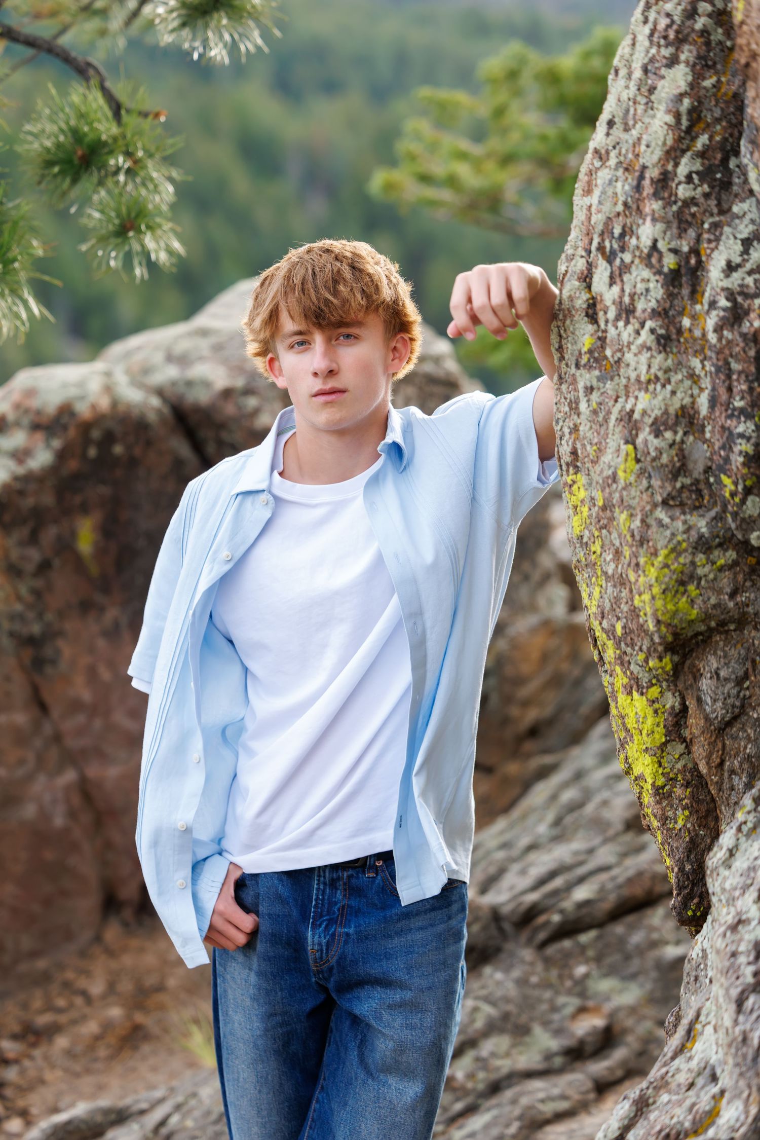 A teenage redhead boy leans against a mossy rock outdoors, wearing a white tee and blue shirt with jeans.