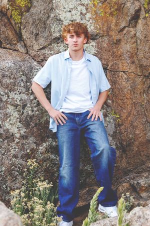 Teen boy in blue jeans and light blue shirt leaning against a rocky wall with wildflowers nearby.