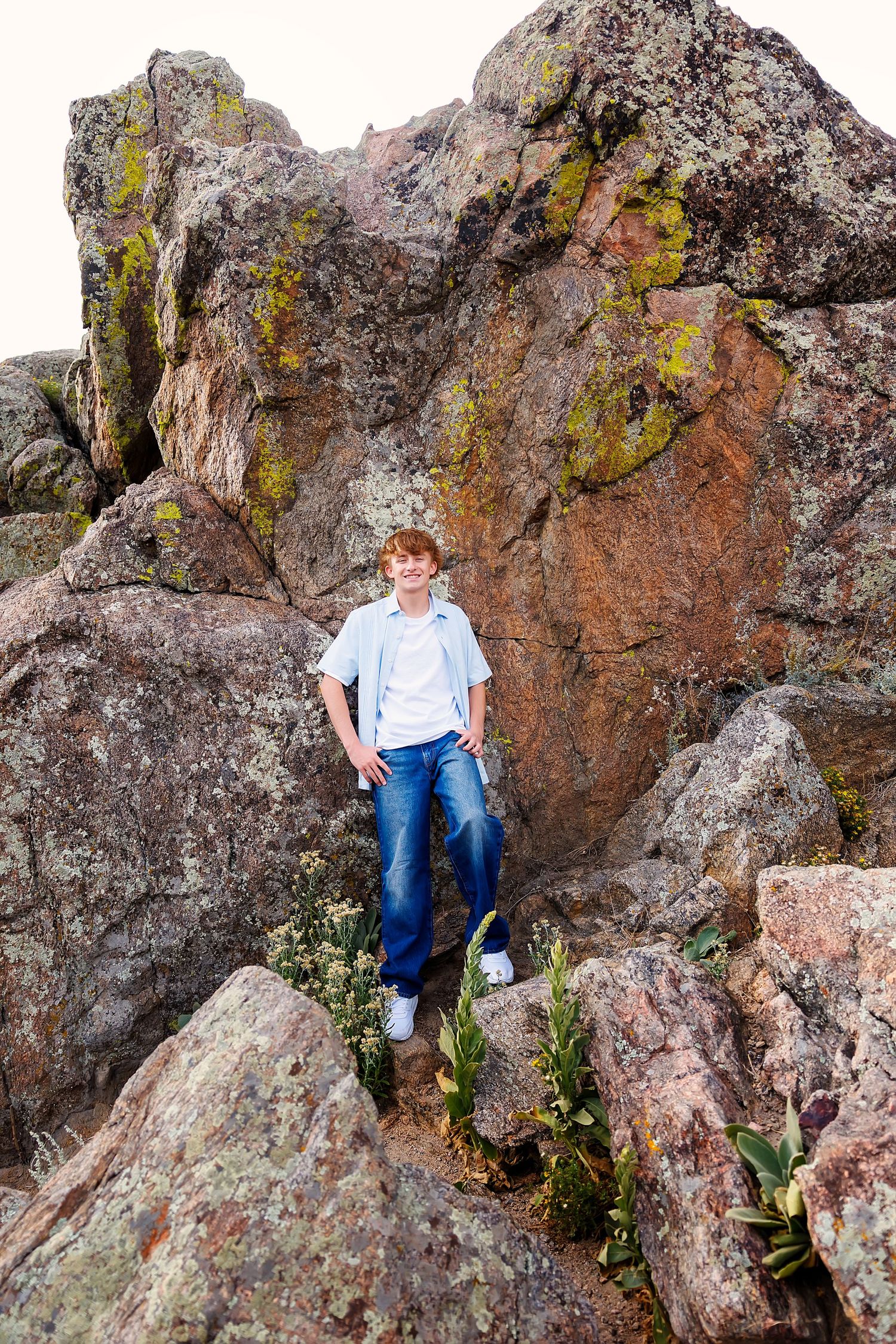 A young man in jeans and blue shirt poses confidently among large moss-covered rocks outdoors.