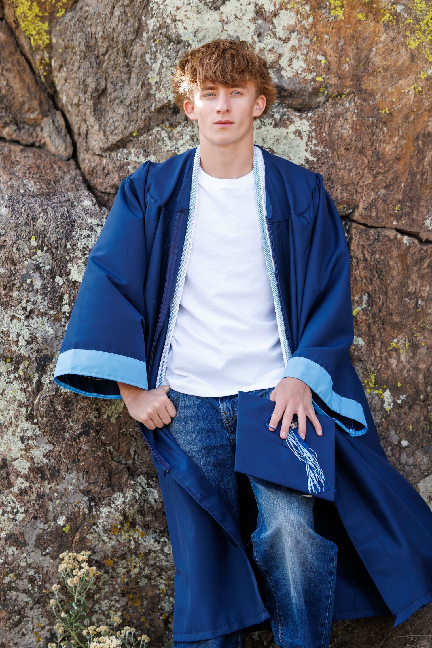 Young man with red hair wearing blue graduation robe over white t-shirt and jeans, leaning against a rock.