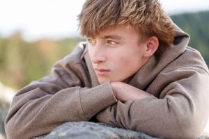 A pensive teenage boy in a brown hoodie rests his chin on crossed arms outdoors with a blurred nature background.