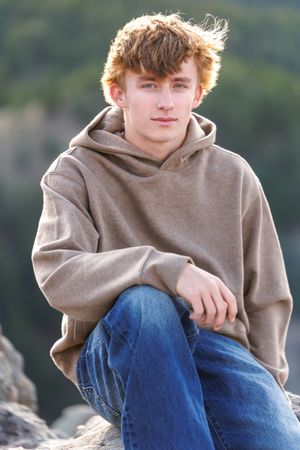 A teenage boy with red hair sits on rocks outdoors, wearing a tan hoodie and jeans, posing for a portrait.