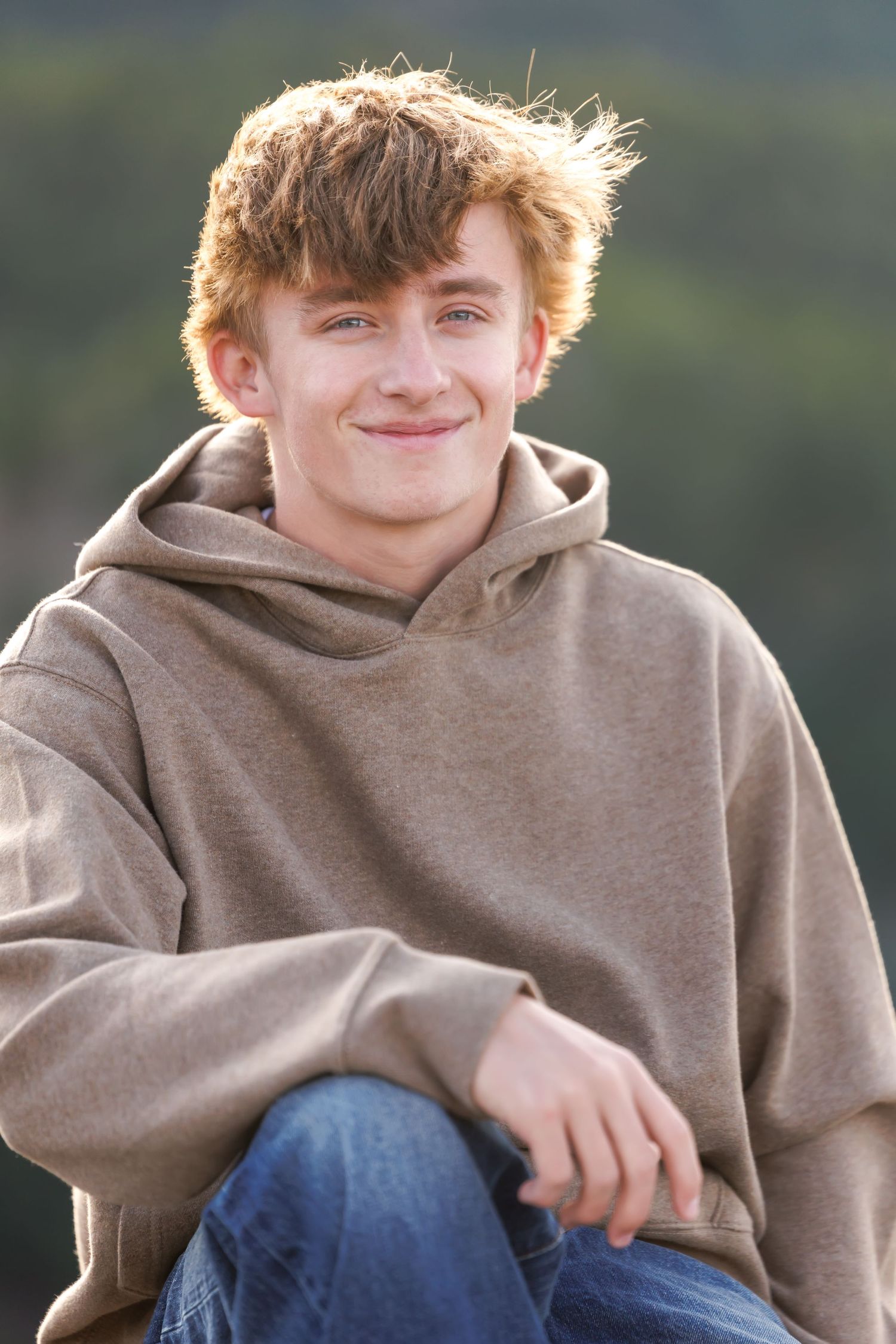 Smiling teenage boy with red hair wearing a brown hoodie and jeans sitting outdoors.
