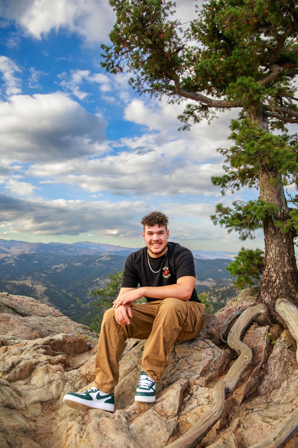 Hiker sits on rocky mountain overlook with scenic mountain vista and blue cloudy sky in background.
