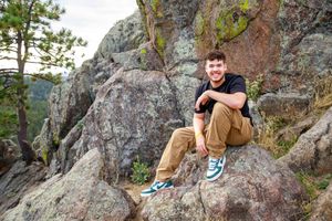 Rock climber in brown jacket scales granite boulder formation in outdoor climbing area.