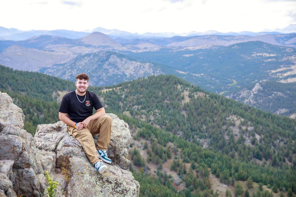 A hiker sits on a rocky mountain summit overlooking a vast forest landscape with rolling hills and mountains in the distance.