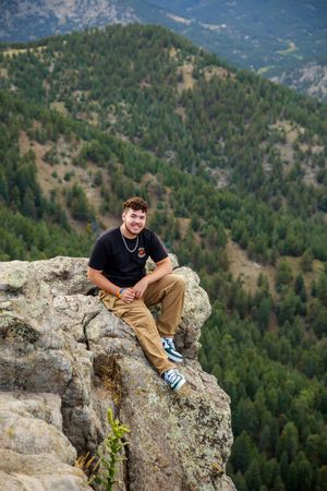 A hiker sits on a rocky cliff edge overlooking a forested mountain valley on a sunny day.