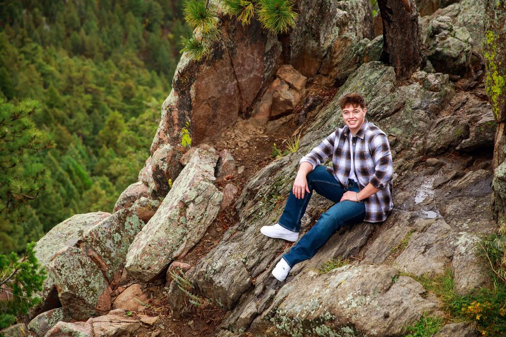 A person in plaid shirt and jeans sits casually on a rocky mountain ledge surrounded by evergreen forest.